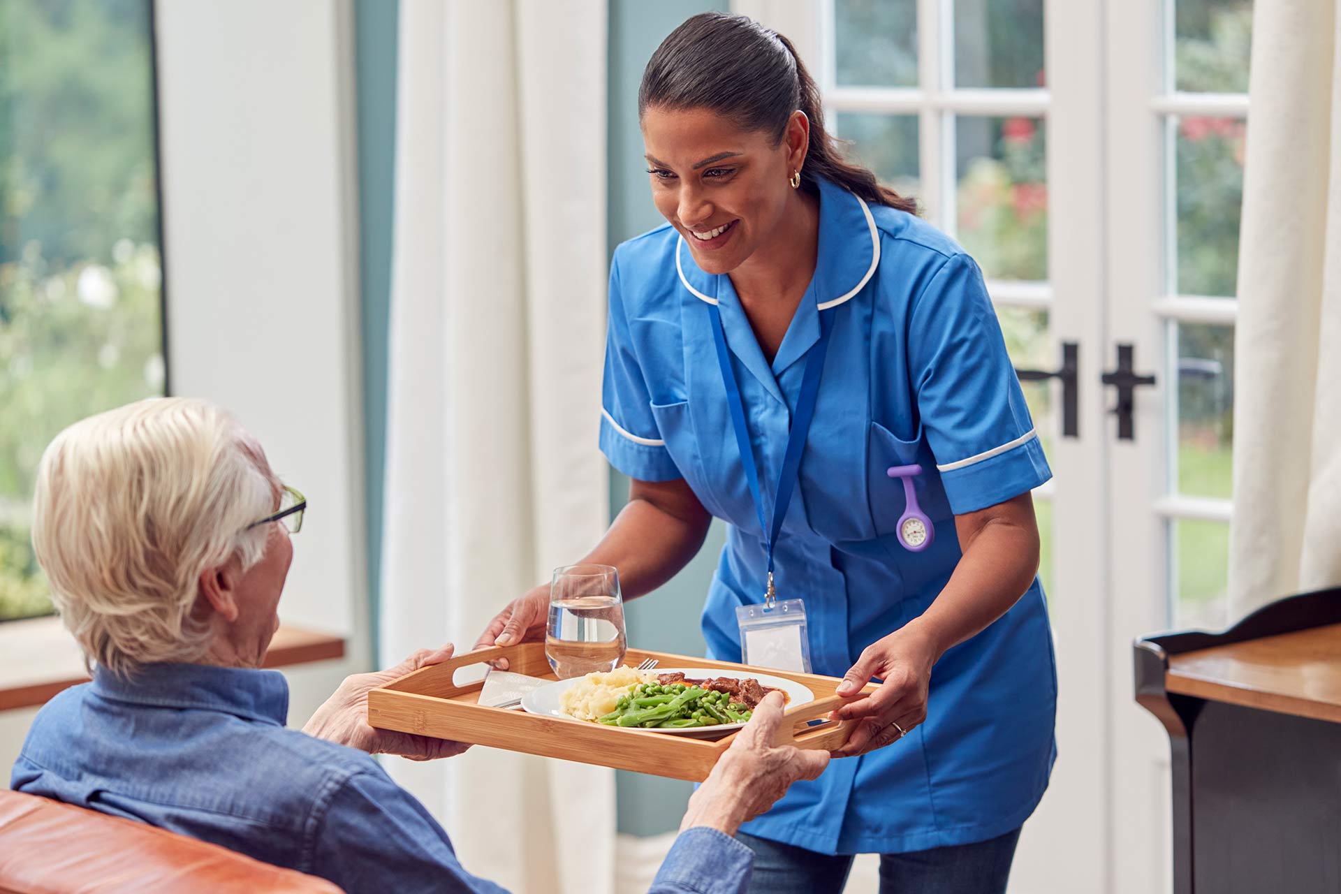 A female carer brings a tray containing a healthy meal and glass of water to a seated elderly person.